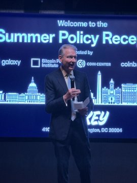 A man in a dark suit speaks into a microphone while holding notes at the "Summer Policy Reception" hosted by Galaxy, Bitcoin Policy Institute, and Coin Center, with the Washington, DC skyline displayed behind him. The image likely captures Hester Peirce delivering her closing remarks emphasizing the importance of preserving the right to transact without an intermediary.