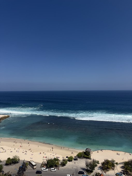 An aerial view of a beach with turquoise ocean waves, a sandy shoreline with people and parked cars, and a clear blue sky.