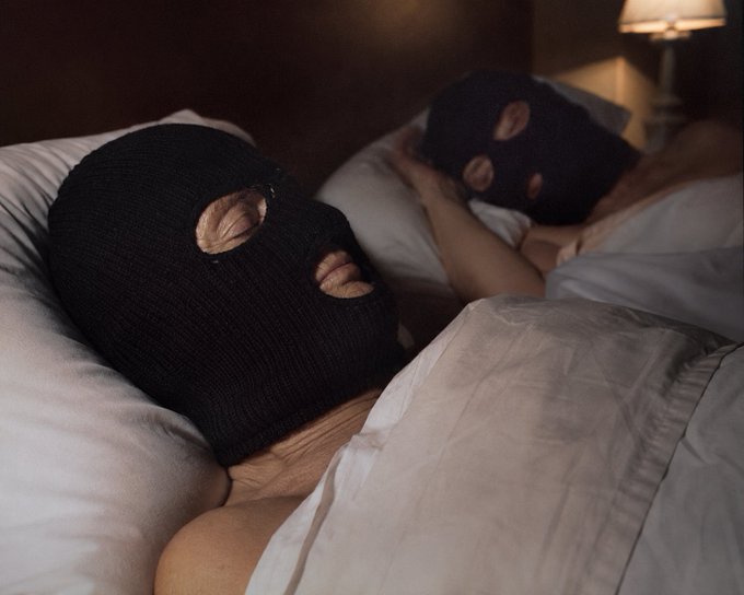 Two people wearing black ski masks lie in bed together under white sheets, illuminated by warm lamplight.