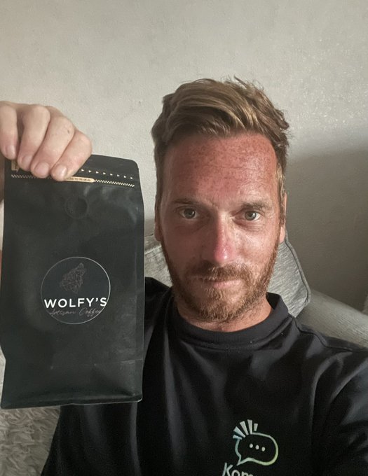 A man holds up a black bag of "Wolfy's Organic Coffee" next to his face, showing off the product with a serious expression. The image accompanies a post about hosting the Wolfy's Bar owner for a steak dinner, with the coffee being the real reason for the invitation, referencing the $KAS ticker.