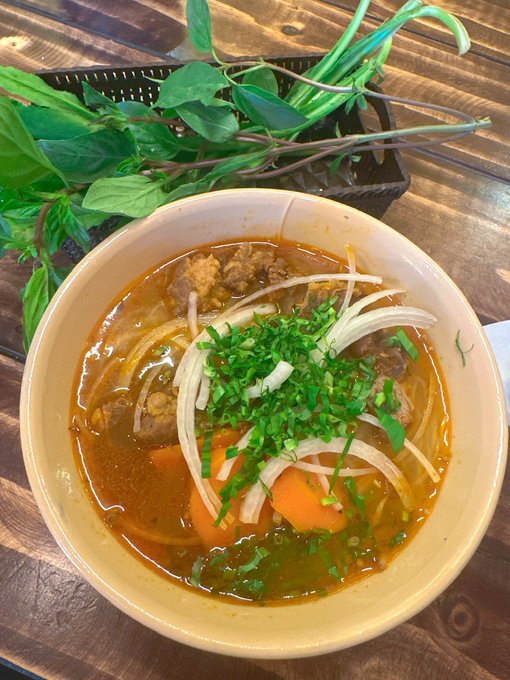 A bowl of reddish broth noodle soup topped with chopped green herbs, sliced onions, tomatoes, and meat, served on a wooden table with fresh basil nearby.