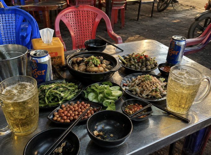A metal table set with a Vietnamese meal including a clay pot of braised meat, green vegetables, cucumber slices, peanuts, chili sauce, and Tiger beer cans alongside frothy mugs of beer.