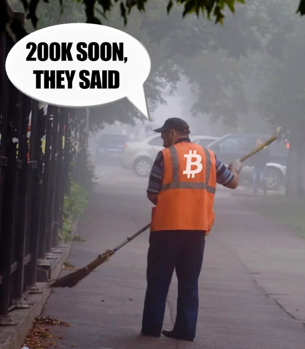 A street sweeper in an orange vest with a Bitcoin logo on the back stands on a foggy sidewalk, with a speech bubble reading '200K SOON, THEY SAID.'