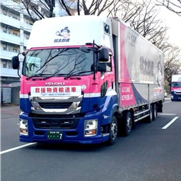 The image shows a blue and pink Isuzu GIGA cargo truck marked "救急物資輸送車" (Emergency Supplies Transport Vehicle) and "AZ-COM Maruwa" operating in Japan, connecting to the announcement about AZ-COM Maruwa HD's billion-yen investment in JPYC, as the company's logistics operations likely facilitate the rapid payment system for partner consignment fees via the dedicated app mentioned.