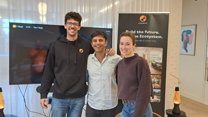 Three people pose together in front of a banner reading 'Build the future. The Ecosystem. You has a London hub' next to a monitor displaying 'Meet 12:39 - Tue 1 Sept'.