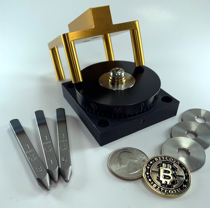 A coin minting press machine with metal stamping dies, displayed alongside a silver coin, a Bitcoin commemorative token, and metal blanks.