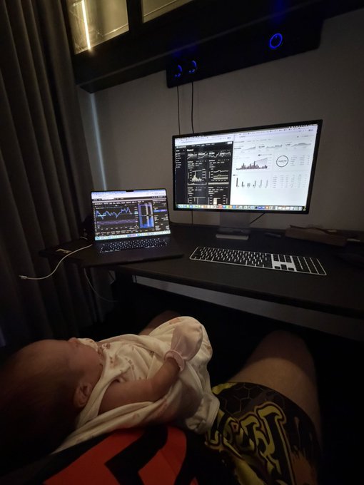 A baby sleeps peacefully on someone's lap in front of a dual-screen workstation, with the laptop displaying stock charts and the monitor showing what appears to be a trading platform with market data, fitting the caption "Got the supervisor sitting in today" as the infant keeps a watchful eye over the trading session.
