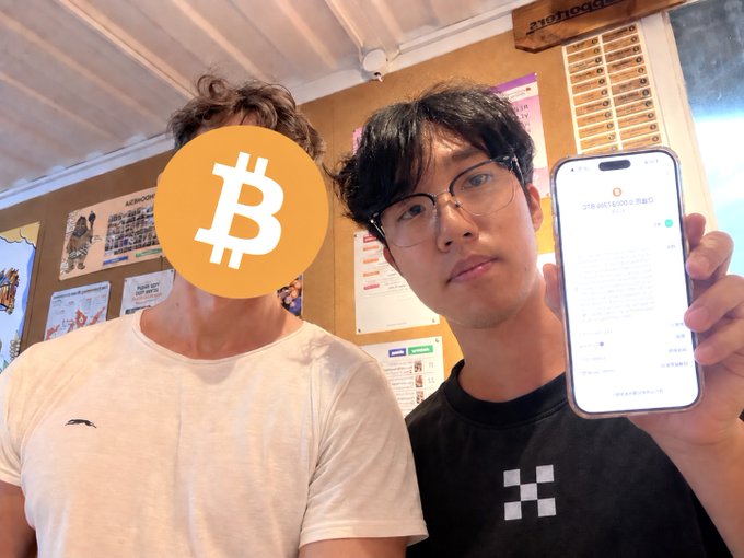 Two young men pose together at Bitcoin House in Bali, with one holding up a smartphone displaying what appears to be a transaction screen (showing text including "320,000" yen/USD and Japanese characters), illustrating their experience visiting this non-profit crypto-accepting café.