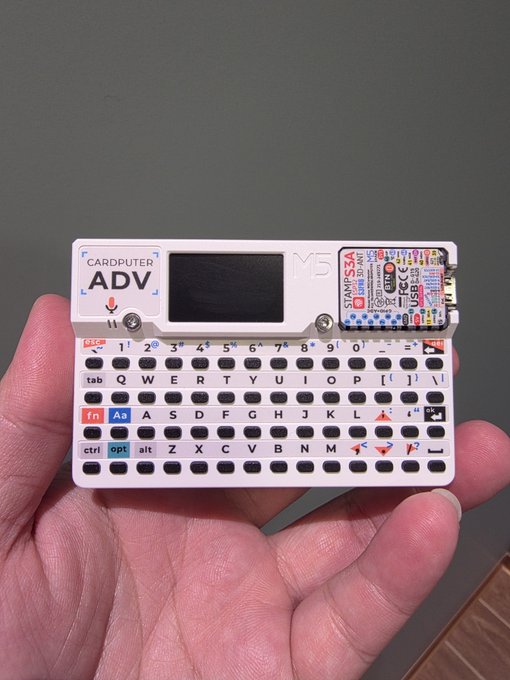 Hand holding a Cardputer ADV V5 mini device with small screen and QWERTY keyboard, featuring a STAMP module on the right side.