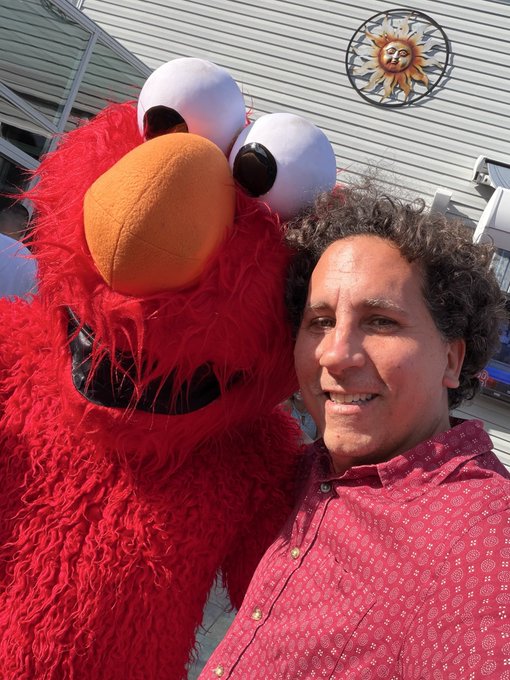 A man poses for a cheerful selfie alongside Elmo, the famous red Sesame Street character, with $DRB noted in the caption as a playful reference to Elmo's well-known enthusiasm. A decorative sun ornament hangs on the light-colored building siding behind them.
