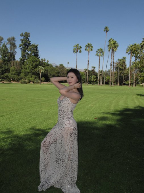 A woman in a long, elegant white and black spotted/sequined gown poses on a manicured green lawn with tall palm trees and mountains in the background under a clear blue sky.