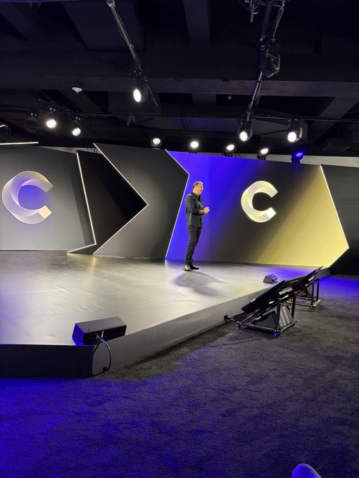 A man stands on a large presentation stage with angular black, blue, and gold panels featuring a stylized 'C' logo, illuminated by overhead spotlights.