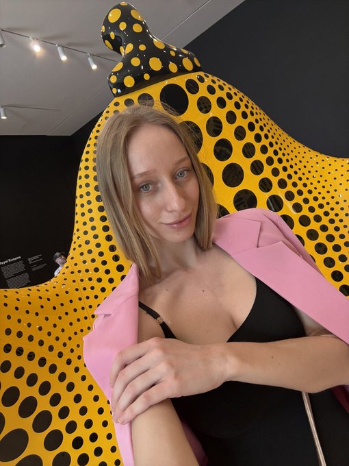 Woman with short blonde hair wearing a pink blazer over a black top, posing in front of a large yellow sculpture with black polka dots in what appears to be an art gallery.