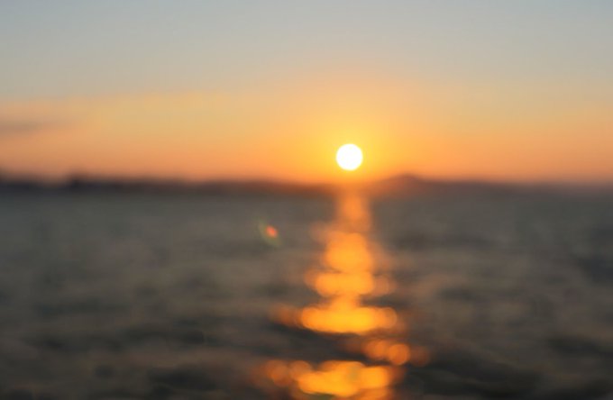 A blurred, dreamy photograph captures a bright orange sunset over a calm body of water, reflecting the sun's glow on the surface.