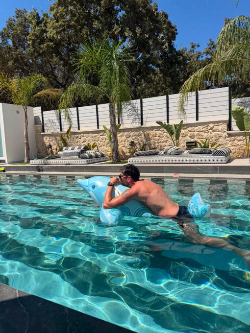 A man wearing goggles swims in a sunny pool while leaning on a blue dolphin-shaped float, with palm trees, loungers, and a stone wall in the background.
