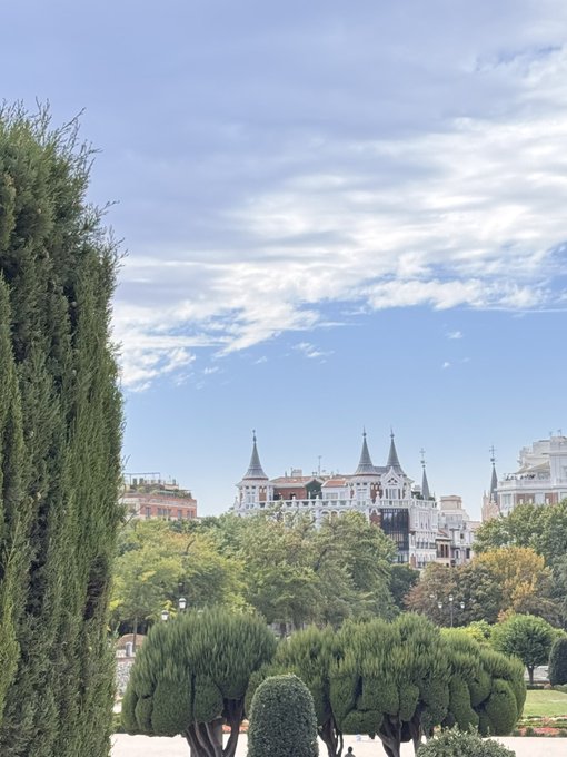 A scenic view shows lush green trees and manicured shrubs in a park, with historic European-style buildings featuring pointed turrets visible in the distance under a partly cloudy blue sky.