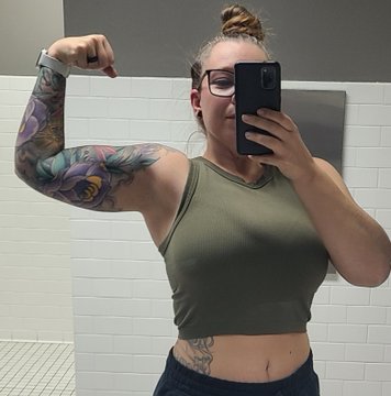 A mirror selfie of a woman wearing glasses and an olive green crop top, flexing her tattooed right arm in front of a tiled wall.