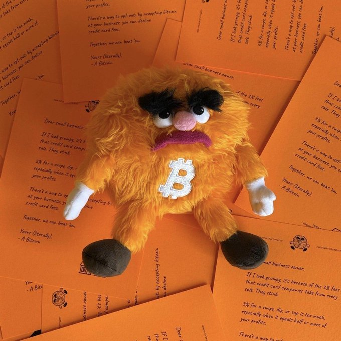 A grumpy orange furry creature with a white “₿” on its chest sits atop orange cards promoting Bitcoin as a way to opt out of credit card fees. The post identifies him as $BITTY, the Bitcoin.org mascot who has appeared on the Satoshi-registered domain since 2015.