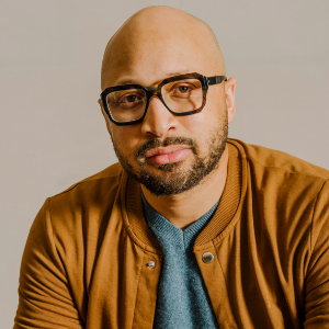 A studio portrait shows a bald man with a short dark beard and mustache wearing black-rimmed glasses, a gray shirt, and a brown bomber jacket, posed against a neutral beige background.