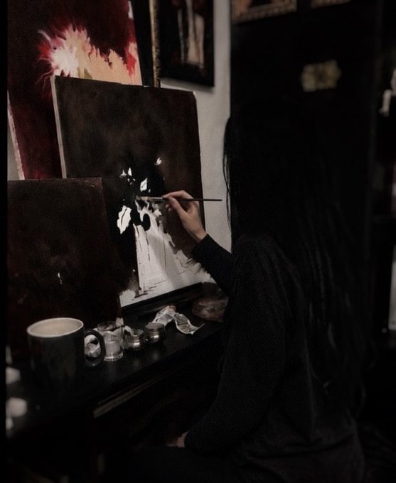 A person with long dark hair paints on a canvas at an easel in a dimly lit room, holding a paintbrush.