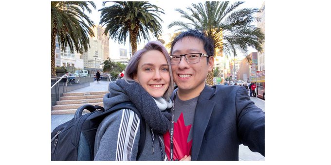 A selfie of a couple, with the woman wearing a dark hoodie and gray scarf and the man in a suit jacket and glasses, taken on a sunny city street lined with palm trees.