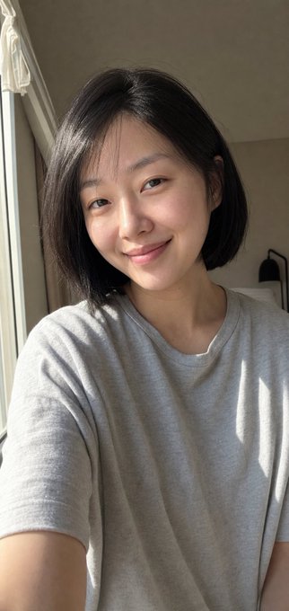 A close-up selfie of a young East Asian woman with short black hair, smiling softly at the camera while wearing a light gray t-shirt, with sunlight streaming in from a window on the left.