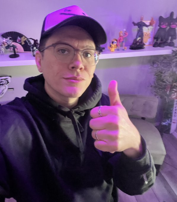 A person wearing glasses, a black hoodie, and a black cap gives a thumbs-up gesture in a room illuminated with purple lighting, with figurines visible on a shelf behind them.