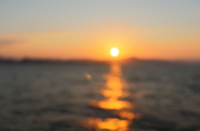 A blurry photograph of a bright yellow sunset over a body of water, with the sun's reflection creating a glowing path on the rippling surface.