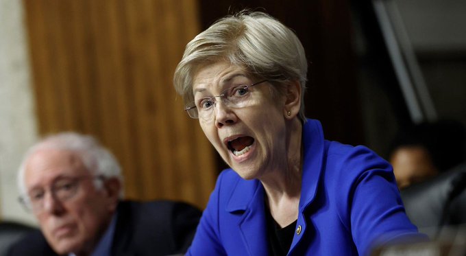 Senator Elizabeth Warren, wearing a blue blazer and glasses, passionately speaks with her mouth open in an animated expression, likely during a hearing where the Clarity Act was being discussed. Her intense demeanor, alongside Senator Bernie Sanders visible in the background, captures the frustration and urgency echoed in the post's reaction to the delayed cryptocurrency legislation vote.