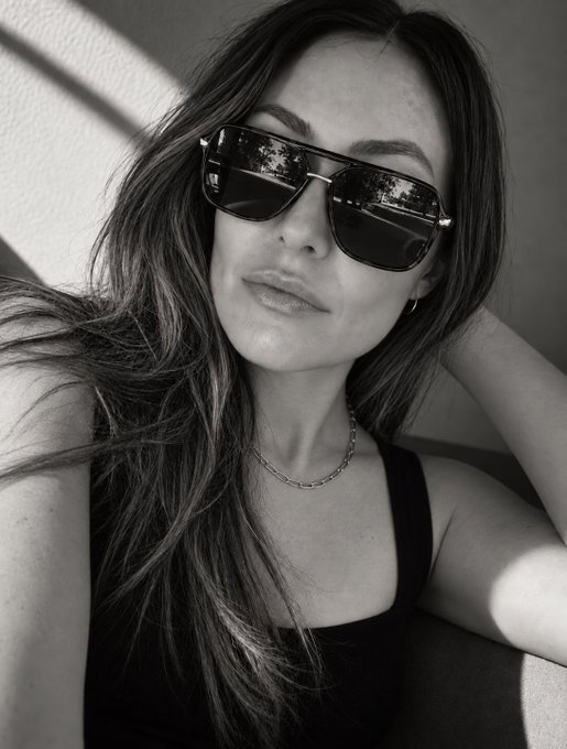 A black-and-white close-up selfie of a woman with long dark hair, wearing sunglasses, a black tank top, and a silver chain necklace.
