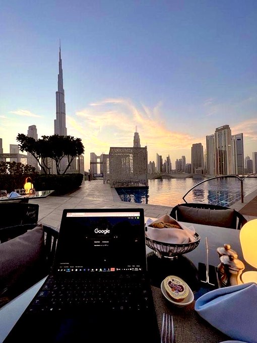 A scenic outdoor view shows a laptop open to Google on a table, with the Burj Khalifa and Dubai skyline visible across the water at sunset.