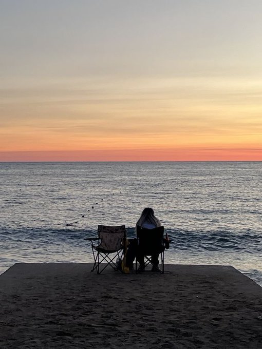 A silhouette of two people sitting in beach chairs facing the ocean during a colorful sunset.