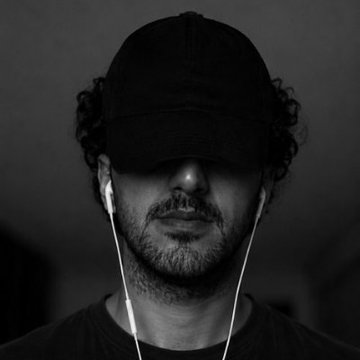 A black and white portrait of a bearded man wearing a black cap and white earphones, with the cap pulled low to obscure his eyes.