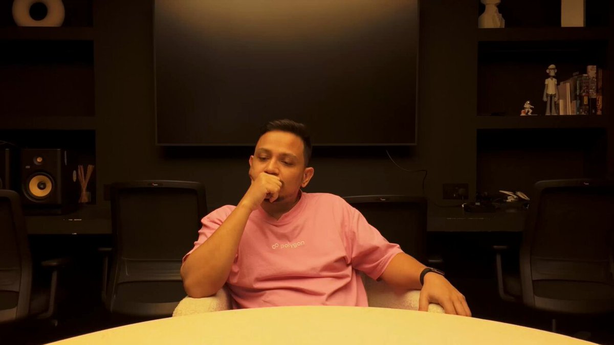 The image shows a man sitting at a table, wearing a pink shirt with the word "Polygon" written on it. The man appears to be deep in thought or contemplation as he looks down into his hand. In front of him are two chairs, one occupied and another empty. A TV screen is visible behind the man, suggesting that this might be an office setting where people work at desks with a television for entertainment purposes.