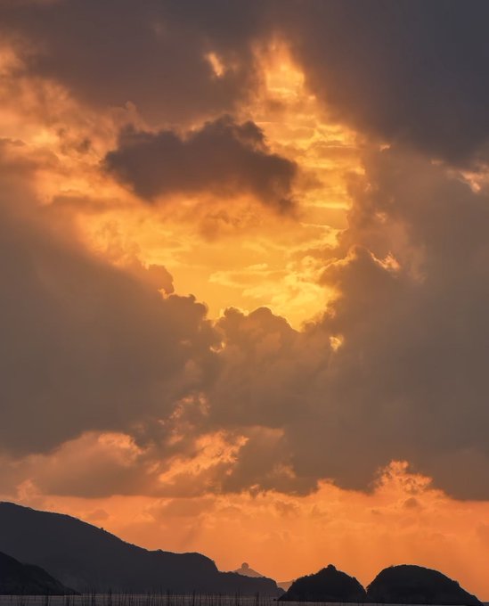 A dramatic sunset photograph featuring dark silhouetted mountains in the foreground beneath a sky filled with voluminous clouds illuminated in rich orange and golden hues.