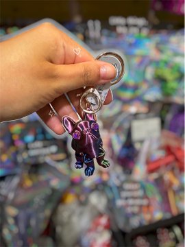 A hand holding a purple and black acrylic keychain charm depicting a stylized character, set against a blurred background of store shelves.