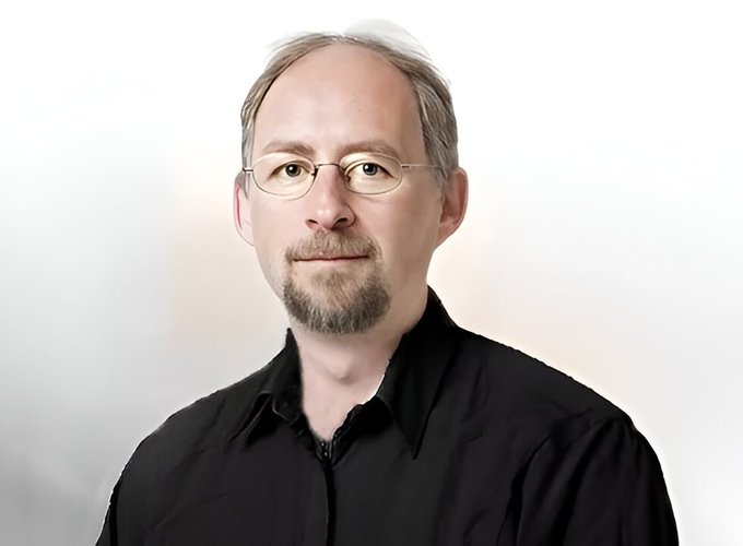 A professional headshot of a middle-aged man with short hair, glasses, and a goatee, wearing a black button-up shirt against a soft, light background.