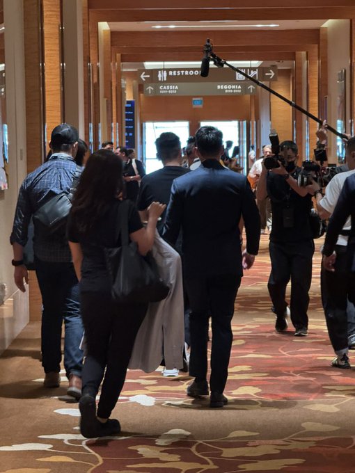A group of people walk down a hotel or conference hallway with patterned carpet, while photographers with cameras and a boom mic follow them.