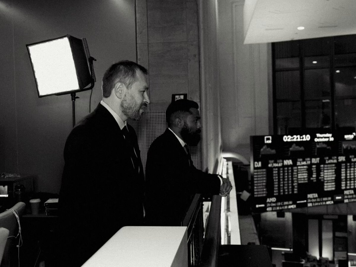 The image shows two men dressed in suits standing next to a large screen, possibly at an airport or train station. They are looking out over the area, which could be a terminal or a waiting room. The focus of the image is on their attire and the surroundings, with no other objects or people visible. There is also a small clock present in the scene, located near the center-left side of the image. The post that reads "the motion chain: Solana" appears to be related to the location where these men are standing, possibly indicating an event or gathering taking place at the airport or train station.