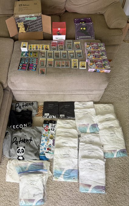 A large collection of VeeFriends and Topps Chrome trading cards is spread out on a couch and floor, including boxes labeled "VeeFriends" and "Topps Chrome," alongside numerous sealed packs, boxes of t-shirts with slogans like "Patient" and "Recon," and items branded with the VeeFriends characters. The image showcases the substantial haul of VeeFriends merchandise the poster is offering in exchange for Bitcoin.