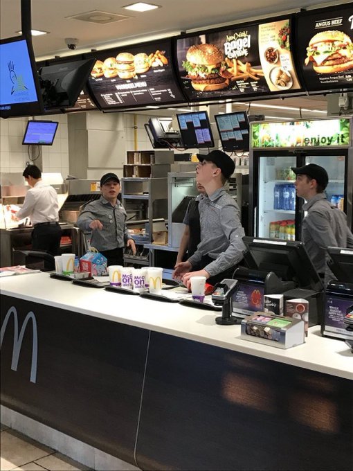 A young man in a McDonald's uniform stands behind the counter beneath menu boards advertising the "Double Cheese" and "Angus Beef" burgers. The image illustrates the post's message about humble beginnings, showing the author's past life earning $3.80/hour before finding success with Bitcoin.