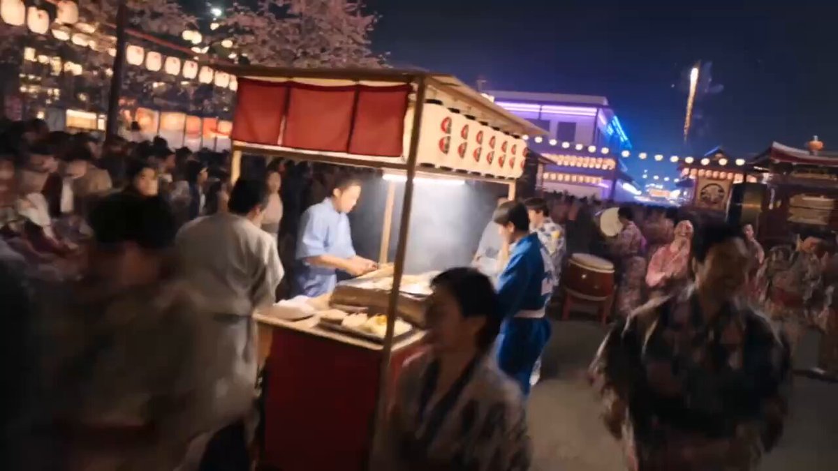 The image shows a lively street scene at night with people walking around and enjoying themselves. There are several food stalls set up along the street, attracting a crowd of people who are eating and socializing. The atmosphere appears to be vibrant and bustling, as if the entire city is alive with energy during this time of day.