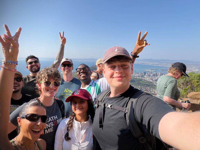 A group of people take a selfie together on a mountain overlook, with some making peace signs and a city and ocean visible in the background.