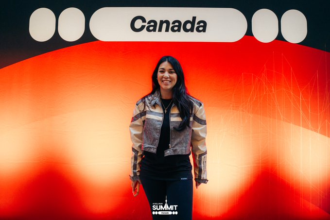 A woman stands smiling in front of a red backdrop displaying 'Canada' at the top, with a 'SUMMIT' logo visible at the bottom center.