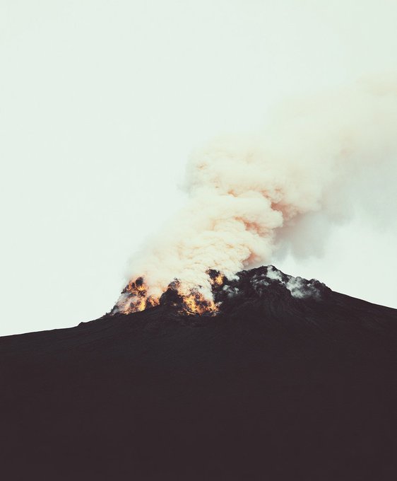 A photograph of a volcano summit erupting with a large plume of smoke and ash rising into a pale sky.