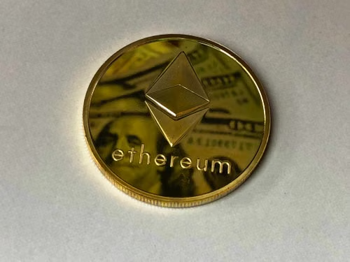 A gold-colored Ethereum (ETH) commemorative coin rests on a white surface, featuring the recognizable diamond-shaped Ethereum logo and the word "ethereum" inscribed below it. The image relates to the post about a significant $37 million ETH withdrawal from Binance, visually emphasizing the cryptocurrency involved in this large-scale transaction.