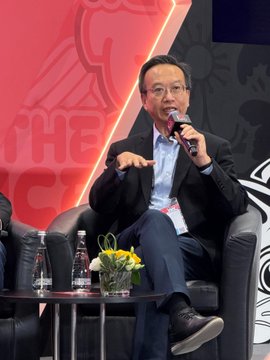 A man wearing glasses and a dark suit speaks into a microphone while seated in a leather chair on a stage with a red backdrop.