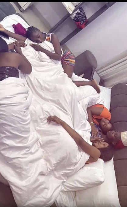 A group of people lie together on a bed under white sheets, appearing to be resting or sleeping in a shared room. The caption "$20 don drop" suggests a humorous or celebratory mood, possibly referencing money or a gift, as the group appears relaxed and comfortable together.