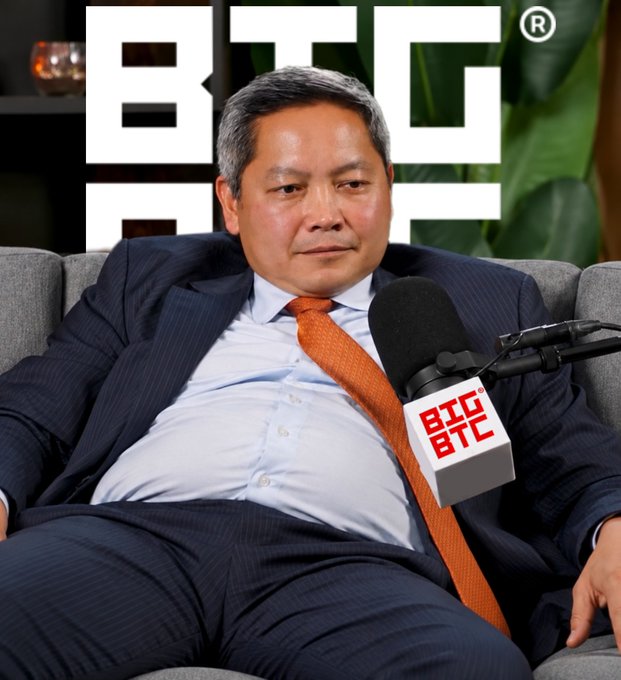 A man in a dark suit and orange tie sits on a gray couch in front of a "BTC" branded backdrop, speaking into a microphone displaying the "BTC" logo, fitting the "Billions Must Comply®" theme of the BTC podcast.