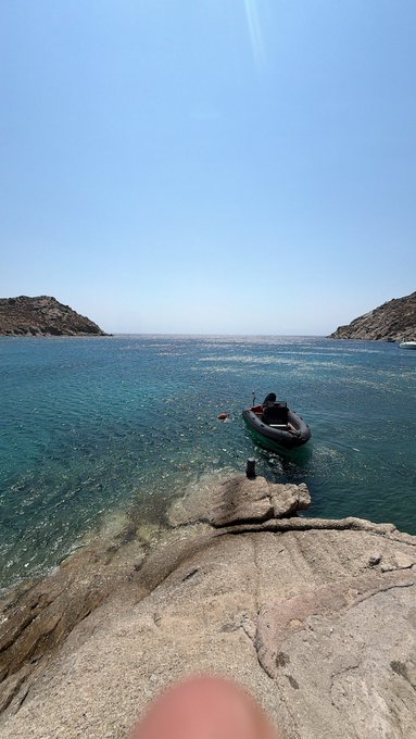 A scenic coastal view showing a person sitting in a small inflatable boat floating in clear turquoise water near rocky cliffs under a bright blue sky, with a finger partially visible at the bottom of the frame.